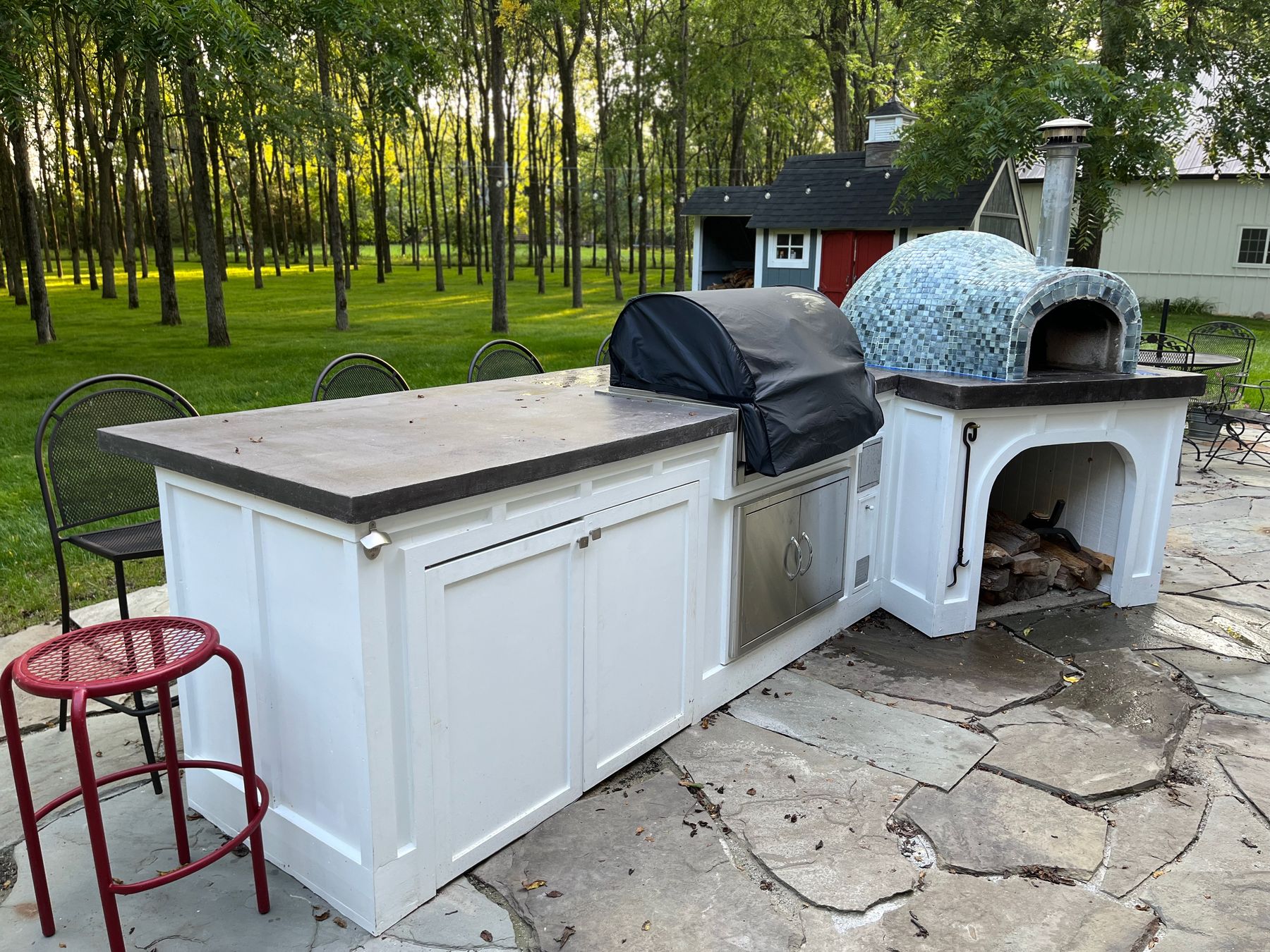 White paneled outdoor kitchen island with a mosaic-tiled pizza oven, set among trees